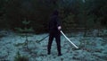 On a dark evening, a man in black ninja clothes stands with his back to the camera and holds a katana in his hand. The