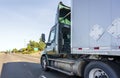 Dark green big rig day cab semi truck with roof spoiler transporting commercial cargo in semi trailer running on the wide highway