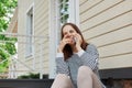 Dark haired adult woman wearing striped shirt and leggins sitting on house porch talking on mobile phone smiling happily having