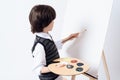 The dark-haired boy draws on the easel. He holds the paint and brush in his hand.