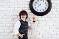 The dark-haired boy is standing by the white wall in a suit. He points at the wall clock. A small future businessman.