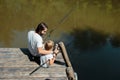 Dark-haired father his little and son dressed in the white t-shirts are sitting with fishing rods on the wooden pier and Royalty Free Stock Photo