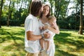 Dark-haired father kisses his beautiful wife holding a small daughter in a white dress in a park on a sunny day. Royalty Free Stock Photo