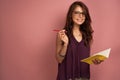 Dark-haired girl in a purple top and glasses with a notebook and pencil stands on a pink background, looks joyfully at Royalty Free Stock Photo