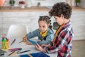 Dark-haired girl sitting at table, writing in her notebook, curly boy standing next to her, showing something on tablet Royalty Free Stock Photo