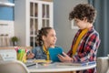 Dark-haired girl sitting at table, writing in her notebook, curly boy standing next to her, showing something on tablet Royalty Free Stock Photo