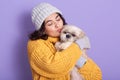 Dark haired girl wearing warm yellow sweater and cap kissing her little dog, blithesome european woman expressing love to her