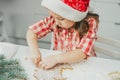 Dark-haired girl 3 years old in red Christmas cap and checkered shirt cuts out gingerbread cookies from rolled dough Royalty Free Stock Photo