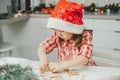 Dark-haired girl 3 years old in red Christmas cap and checkered shirt cuts out gingerbread cookies from rolled dough Royalty Free Stock Photo