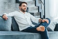 Dark-haired handsome, barefoot confident, smiling bearded businessman sitting on soft blue couch in modern living room