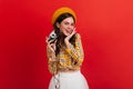 Dark-haired lady in orange beret is holding retro camera. Girl in bright blouse smiles coquettishly on red background. Royalty Free Stock Photo