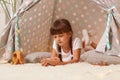 Dark haired little girl with braids wearing white t shirt posing in peetee tent, lying with cell phone, using smart phone for Royalty Free Stock Photo