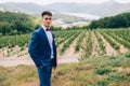 The dark-haired man in a blue suit walks in nature and poses against the background of green vineyards, mountains and Royalty Free Stock Photo