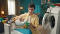 Dark-haired man sitting next to a washing machine, pouring a liquid detergent into a cup to fill the dispenser, planning Royalty Free Stock Photo