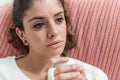 Dark-haired, thoughtful young woman takes a cup sitting on the couch.