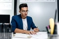 Dark-haired young architect in glasses and in a blue jacket is working with documents on the desk in the office Royalty Free Stock Photo