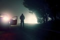 A dark moody concept of a threatening hooded figure standing next to a parked car. On a city street at night