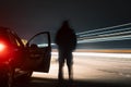 A dark mysterious blurred figure standing on a road, next to car. Next to traffic trails on a country road at night Royalty Free Stock Photo