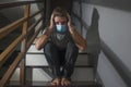 dark portrait of young scared and worried man in protective mask sitting on stairs at home staircase during lockdown quarantine Royalty Free Stock Photo