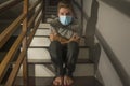 Dark portrait of young scared and worried man in protective mask sitting on stairs at home staircase during lockdown and Royalty Free Stock Photo