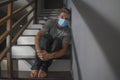 Dark portrait of young scared and worried man in protective mask sitting on stairs at home staircase during lockdown and Royalty Free Stock Photo