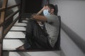 Dark portrait of young scared and worried man in protective mask sitting on stairs at home staircase during lockdown and Royalty Free Stock Photo