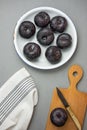 Dark red big plums on white vintage enamel plate. Wooden cutting board knife white cotton towel on gray stone. Minimalist