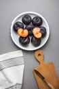 Dark red big plums whole and halved on white vintage enamel plate. Wooden cutting board knife white cotton towel on gray stone