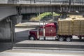 Dark red big rig classic semi Truck with flat bed semi trailer carries pressed hay running on the highway under the bridge Royalty Free Stock Photo