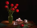 In a dark room, on a wooden table is a green vase with tulips. next to the table are two white cups and a plate of marshmallows Royalty Free Stock Photo