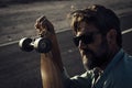 Dark shadow and light portrait of handsome adult man with beard and sunglasses holding long board with asphalt road in background