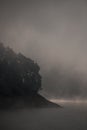 dark silhouette of a hillside with trees near the water covered with white haze and fog Royalty Free Stock Photo