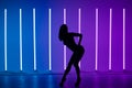 A dark silhouette of a slender sexy young woman posing high heels against a background of neon lamps.