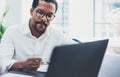 Dark skinned coworker wearing glasses and using laptop in modern office.African american man in white shirt working on