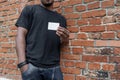 Dark-skinned man in T-shirt showing blank business card on bricked background. Royalty Free Stock Photo