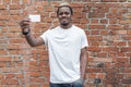 Dark-skinned man in white T-shirt showing business card on bricked background. Royalty Free Stock Photo