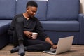 Dark skinned young male working with his laptop at home, holds cup with hot beverage, sitting near couch on floor, handsome man Royalty Free Stock Photo