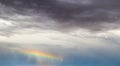 Dark stormy sky with horizontal rainbow in blue sky in distance with rain falling out of it