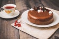 On a dark tabletop, on a linen napkin on a white dish, there is a round chocolate cake, next to a white cup is dark tea and next