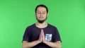 A darkhaired young man in a black Tshirt holds his palms together in a prayer gesture. A man in the studio on a green