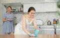 Daughter dusting table while mother washes dishes in kitchen