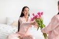 Daughter gives flowers to smiling pretty asian young woman in pink pajama on bed in bedroom interior Royalty Free Stock Photo