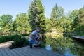Dawn. A young man squats on the edge of a wooden deck in front of a small picturesque pond and looks down at the water