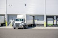 Day cab white big rig semi truck with dry van semi trailer standing on the warehouse dock gate loading cargo for the next Royalty Free Stock Photo