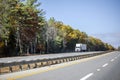 Day cab white big rig semi truck transporting cargo in container trailer running on the divided highway with autumn trees line Royalty Free Stock Photo