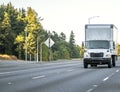 Day cab white middle powered semi truck with box trailer running on the highway road with local entrance and autumn trees on the Royalty Free Stock Photo