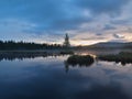 Daybreak autumn lake with mirror water level in mysterious forest, young tree on island in middle. Fresh green color of herbs