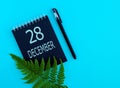 December 28th. Day 28 of month, Calendar date. Black notepad sheet, pen, fern twig, on a blue background