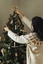 Decorating christmas tree with stylish golden star and ribbon. Woman in cozy sweater hanging vintage ornament on tree branch close Royalty Free Stock Photo