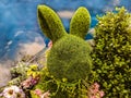 Decorative green topiary rabbit head with flowers around its neck and surrounded by greenery in front of blue sky-like background